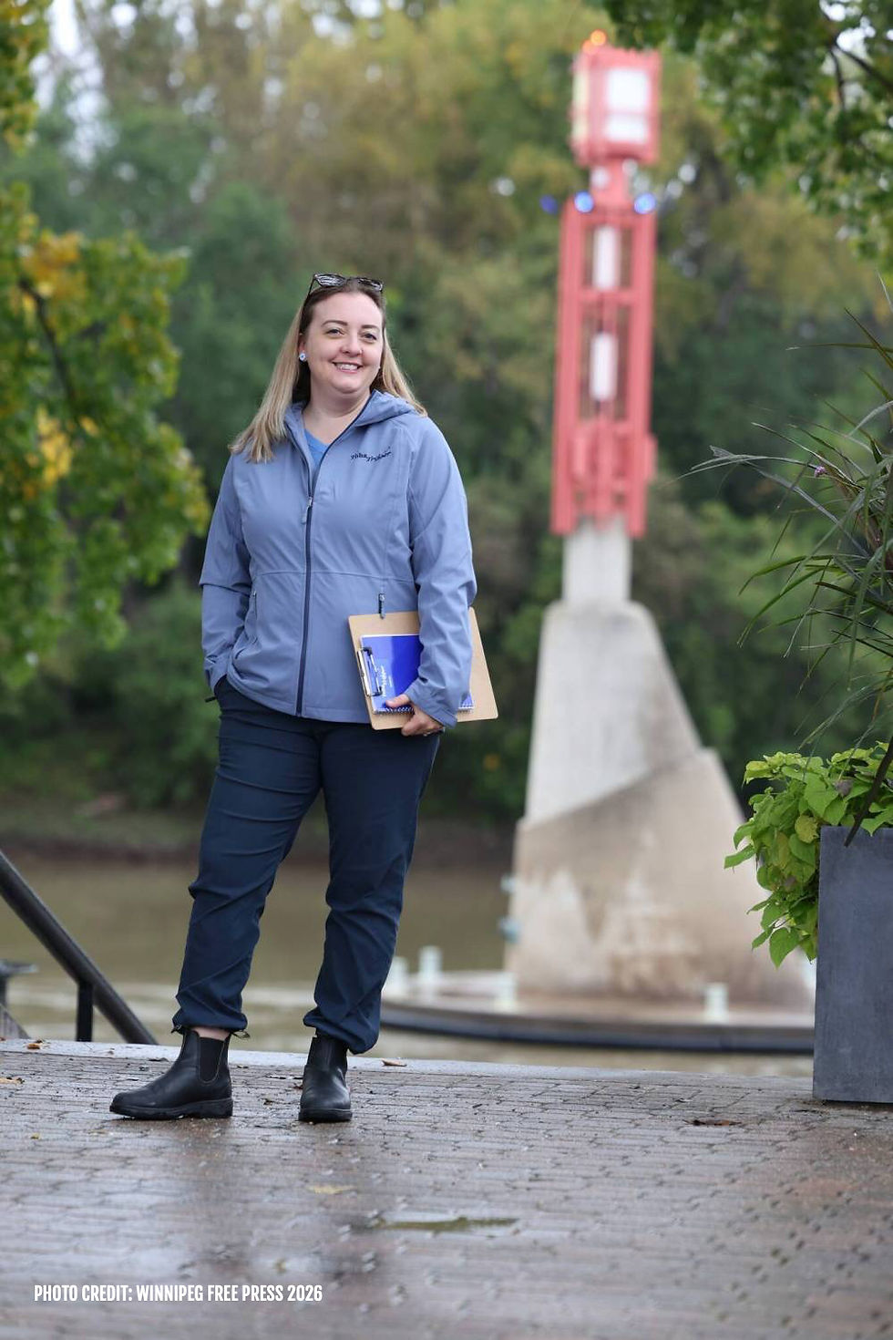 Woman with blonde hair standing outside in a light blue coat holding a clipboard.