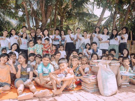 A group of smiling children and adults outdoors under trees, posing with books and a sack. They make heart gestures on a sunny day.