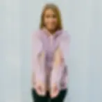 Woman in a pink hoodie shows off zippered sleeves, smiling against a light-colored wall. Relaxed mood.