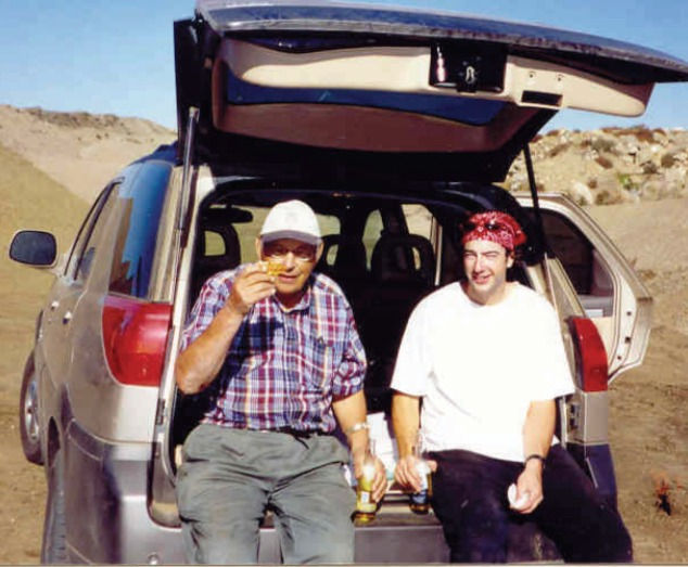 two men sit on the open tailgate of an SUV, surrounded by sand. One is old, and one is young