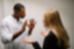 Man and woman engaged in a serious discussion indoors, gesturing with hands. He wears a white shirt, she wears black. Neutral background.