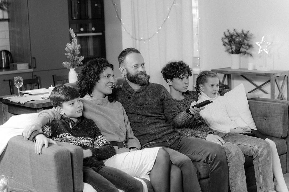 Family of five sits on a couch in a cozy living room, watching TV. Warm lighting, fairy lights, and a small tree set a festive mood.