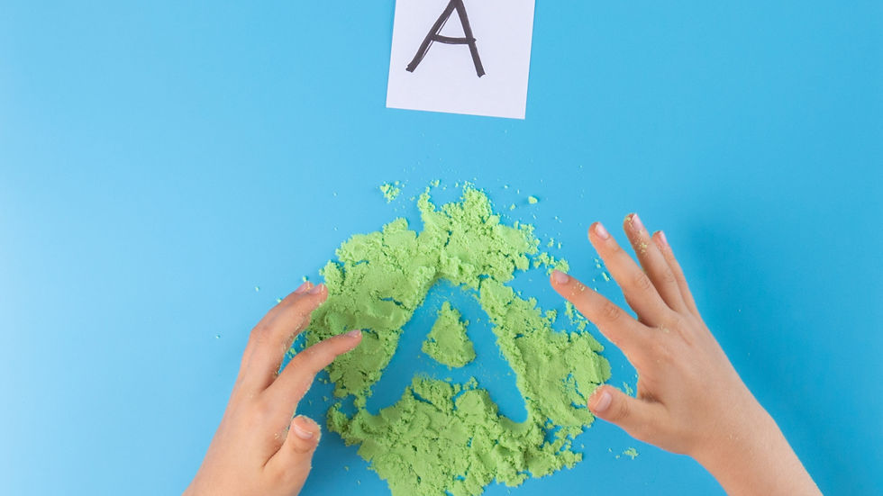 Child's hands shape a green "A" in sand on a blue surface. A paper with "A" is above, evoking a playful, learning mood.