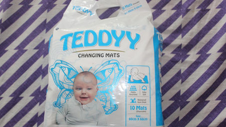 Packaging of TEDDYY changing mats with a smiling baby, butterfly design, and product details on a purple patterned background.