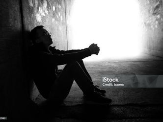 Silhouetted man sits curled up in a concrete tunnel, facing bright light; iStock Credit: baona and 2149642583 visible.