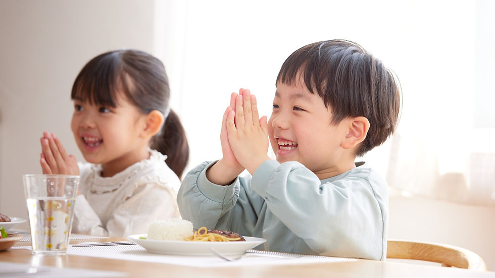 Two smiling children sit at a table, hands together in a grateful gesture. Plates of food in front, bright daylight fills the room.