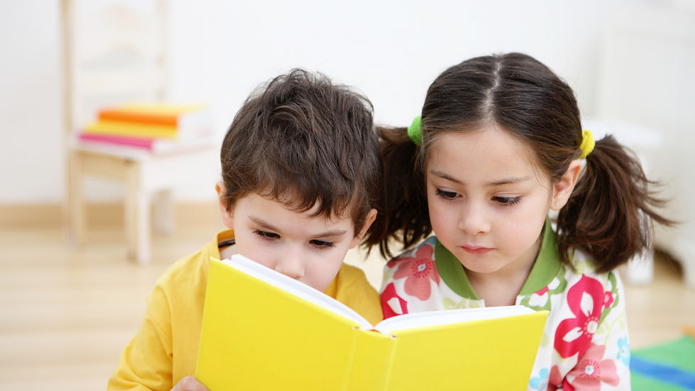 Two children intently read a bright yellow book. One wears yellow, the other floral. Soft background with a stack of books.