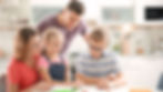 Family of four in a bright kitchen, smiling and collaborating on homework. The girl writes as parents and brother look on approvingly.