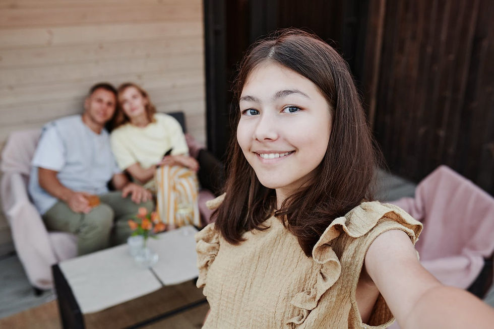 Teen girl takes a selfie with a smile. In the background, two adults sit on a patio couch. Light wood walls and cozy setup.