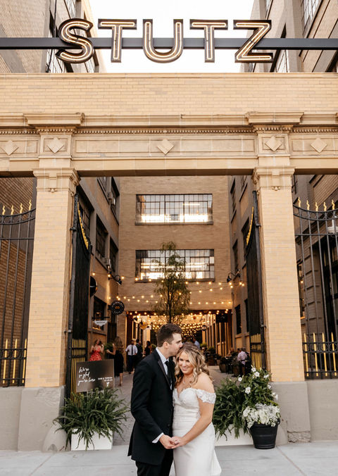 Groom kissing bride's cheek in front of facade of Stutz