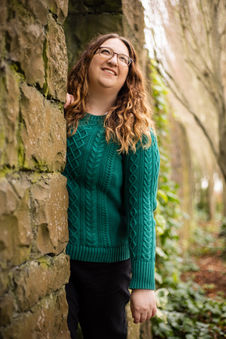 Shauna Carrick, wearing a green knitted jumper, is leaning out from behind a stone wall, looking upwards to the sky.