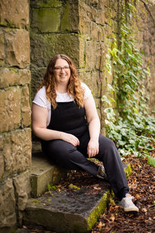 Shauna Carrick, wearing a white short sleeved t-shirt and a pair of black dungarees, sits on a stone step, smiling at the camera. She looks to be in a wooded area, with greenery visible behind her.