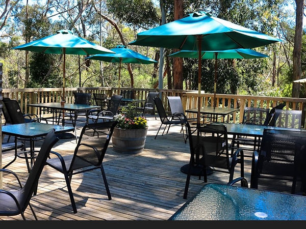 Outdoor timber deck seating area surrounded by bushland, featuring glass-top tables, black chairs, and green umbrellas, with
