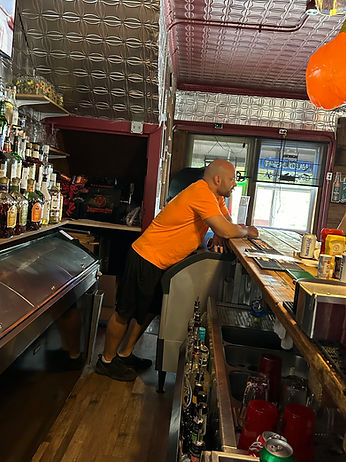 Bald man in orange shirt leans against a dimly lit bar.