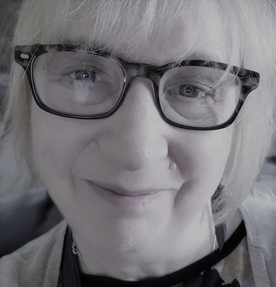 Person with glasses smiling softly, close-up portrait, light hair, black frames, neutral background, conveying a calm, friendly mood.