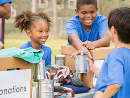 Children in blue shirts volunteer, smiling and handing out canned goods at a donation table outdoors. Boxes labeled "donations" are visible.