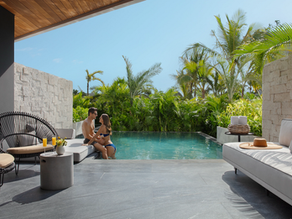 Couple in swimsuits relax by a pool in a tropical setting, surrounded by palm trees. Modern patio furniture and drinks in foreground.