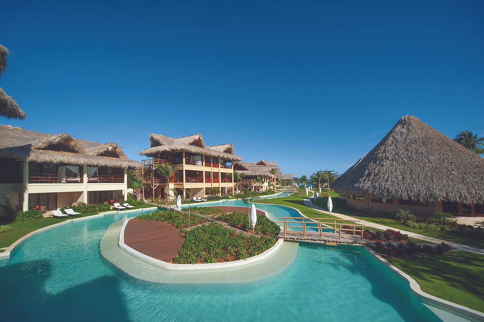 Tropical resort with huts, a winding pool, and lush greenery under a clear blue sky. Relaxed, inviting atmosphere.