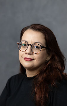 mulher braca, com cabelos ruivos escuros compridos, óculos de tartaruga, em frente a uma parede de cimento queimado.