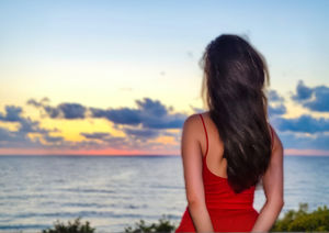 A woman in a red dress, standing at the edge of the day, letting the ocean carry her thoughts into the sunset.