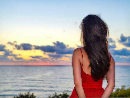 A woman in a red dress, standing at the edge of the day, letting the ocean carry her thoughts into the sunset.
