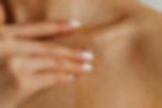 Hand with white nails gently touching a shoulder. Skin appears hydrated, with a warm, neutral background, evoking a calm, soo
