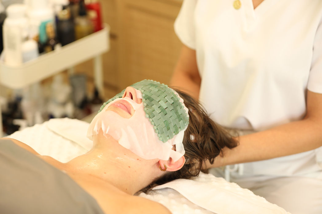 A person receives a facial treatment with a green mask at Transcendence Spa, while Marcia, the aesthetician, attends to them in a calm setting.