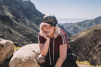 Blonde white actress, Emilia Ray, on a moutain, looks at the camrea with a hat and wearing a t-shirt