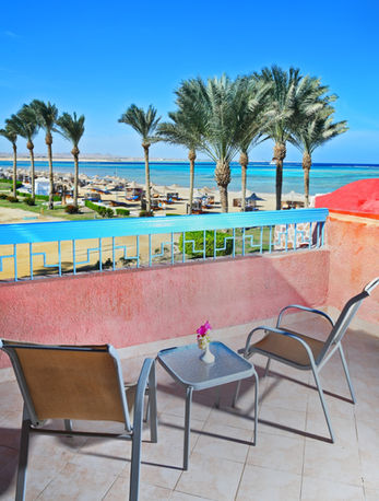 Beach view from balcony with chairs and table, palm trees, blue water.