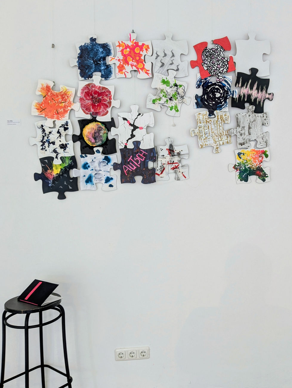 Foto des Puzzles mit Lücken und 19 bemalten Puzzlestücken. Ein Puzzlestück bleibt weiß. Davor ein Barhocker auf dem das Heft liegt.