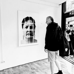Besucher steht for dem Portrait (UN)GESEHEN in der Ausstellung KOPFLANDSCHAFTEN. Das Portrait zeigt das Gesicht der Künstlerin in Nahaufnahme, mit verschwitzten Haaren, Schweißtropfen und großen kettenförmigen pinken Ohrringen. Das Foto ist in schwarz-weiß.