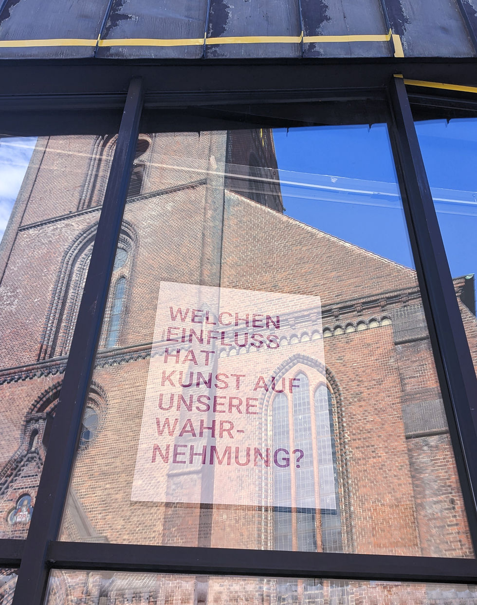 Foto eines Fensters. Darin spiegelt sich eine Kirche im Klinkerstil. Im Fenster hängt ein Poster auf dem die Frage steht: Welchen Einfluss hat Kunst auf unsere Wahrnehmung?