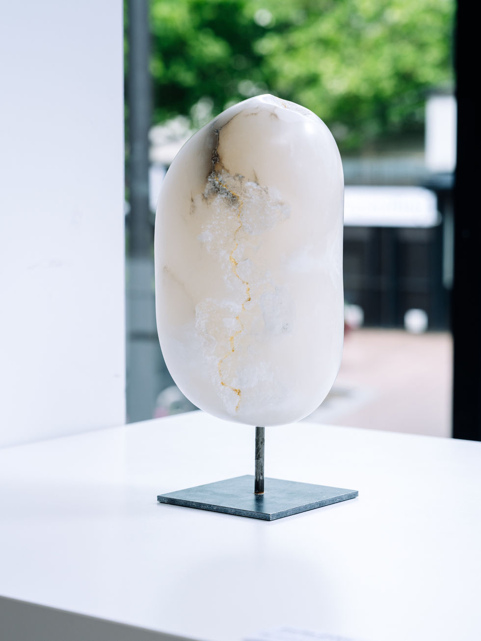 The sculpture stands on a white pedestal. In the background, a square with trees can be seen through a window.