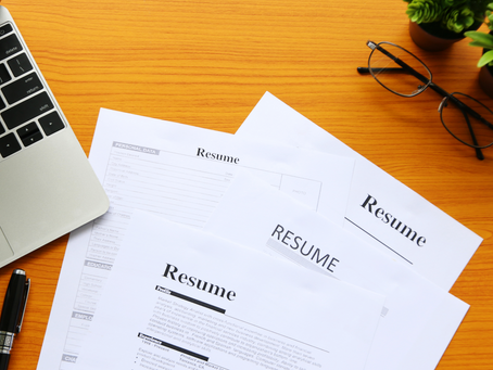 A stack of resumes scattered on a desk with a laptop, pen, eye glasses, and two small plants.