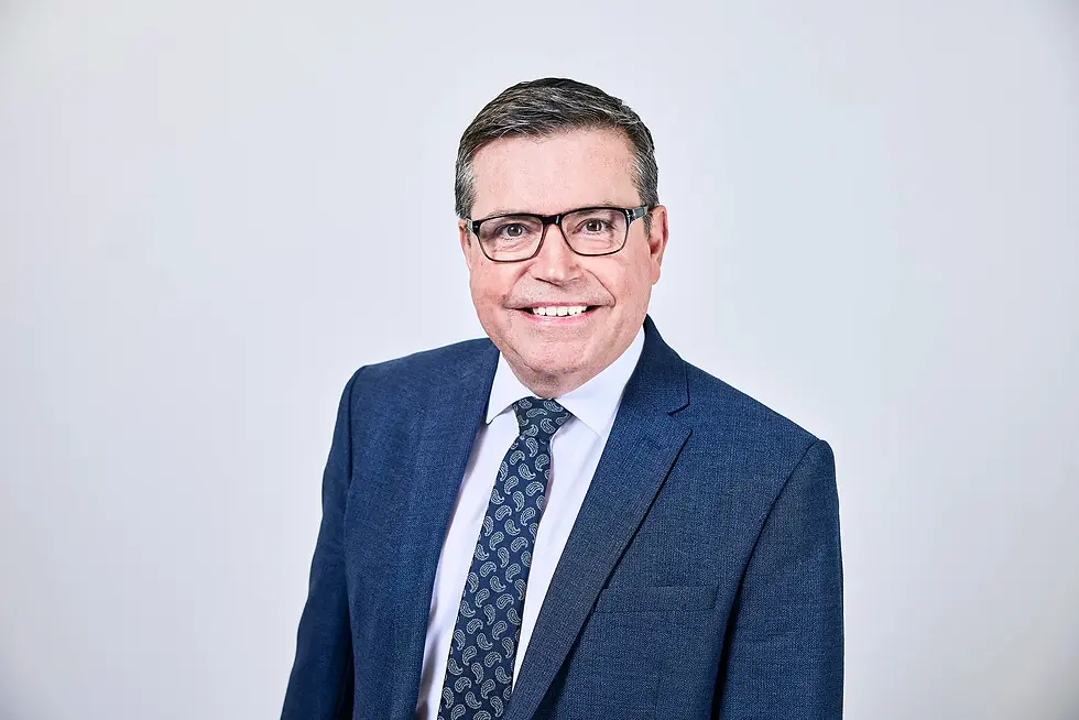 Professional headshot of Tim Foreman smiling, wearing glasses, a blue textured suit jacket, a white dress shirt, and a patterned dark blue tie against a plain light grey background.
