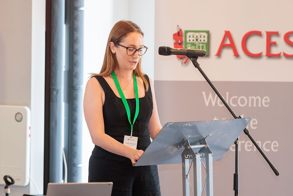 Sophie Davidson standing at a lectern reading notes behind a microphone with an ACES banner in the background.