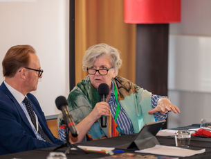 Jackie Sadek speaking into a microphone while seated at a conference panel table next to a male colleague.