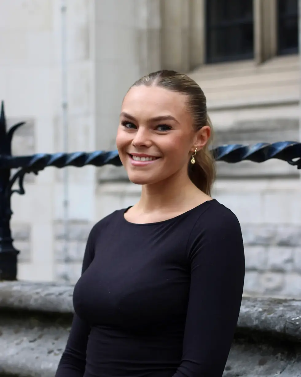 Professional headshot of Grace Esberger smiling, with her hair tied back in a sleek ponytail, wearing a black long-sleeved top and gold drop earrings, outdoors against a blurred stone building and iron railing.