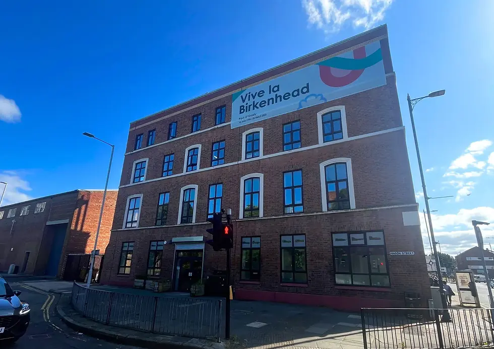 A three-story red brick commercial building on a street corner featuring a large white banner that reads "Vive la Birkenhead".