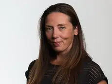 Professional headshot of Andrea Jutrzenka smiling faintly, with long straight brown hair, wearing a black top with horizontal sheer stripes against a plain white background.