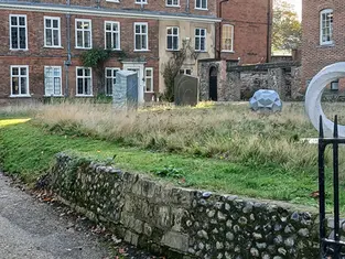 A historic red brick office building with white gabled trim in The Close, Norwich, featuring a flint stone wall and modern sculptures in the front garden.