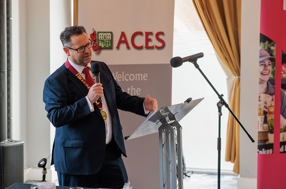 ACES President Alan Richards wearing the presidential chain of office and speaking into a handheld microphone at a lectern.