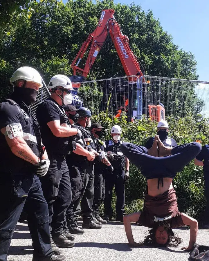 A protester performing a tripod headstand with legs in a cross position on a road, directly in front of a line of enforcement officers and a large red Hitachi excavator.