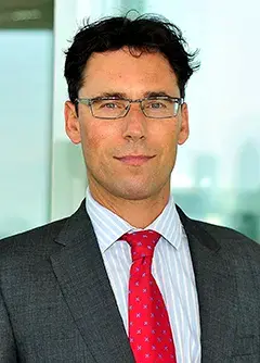 Professional corporate headshot of Guy Brett smiling slightly, wearing glasses, a grey suit jacket, a striped shirt, and a red tie.