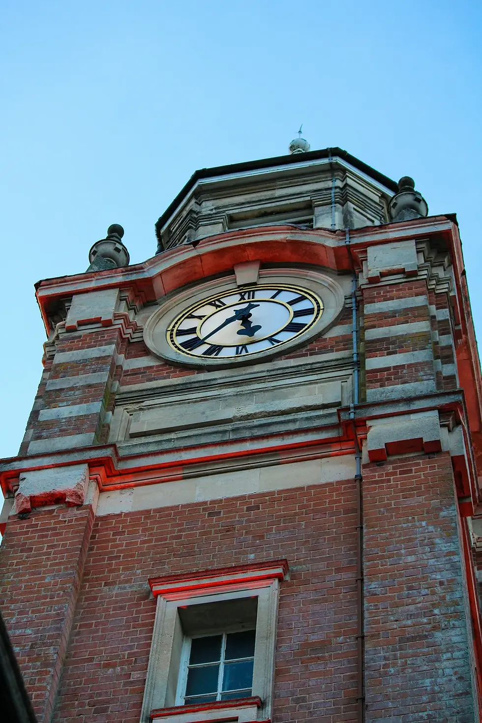 Low-angle shot looking up at a brick and stone clock tower with Roman numerals against a clear blue sky.