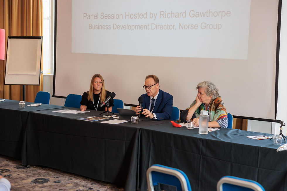 Richard Gawthorpe speaking into a microphone while seated between Lauren Stretch and Jackie Sadek during the Norse Group housing delivery panel session.