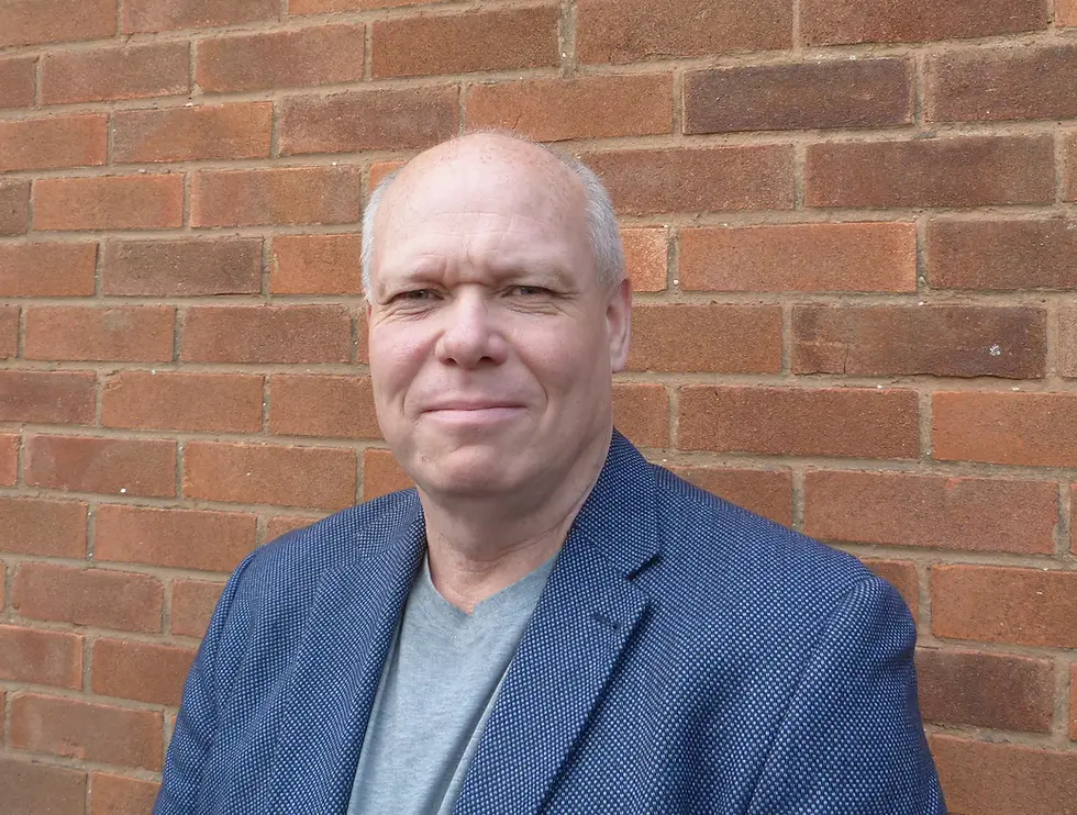 Professional headshot of Phil Farrell smiling faintly, wearing a blue patterned textured blazer and a grey v-neck t-shirt, standing in front of a red brick wall.