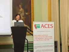 Woman speaking at a conference podium with an ACES (The Association of Chief Estates Surveyors and Property Managers in the Public Sector) banner visible.