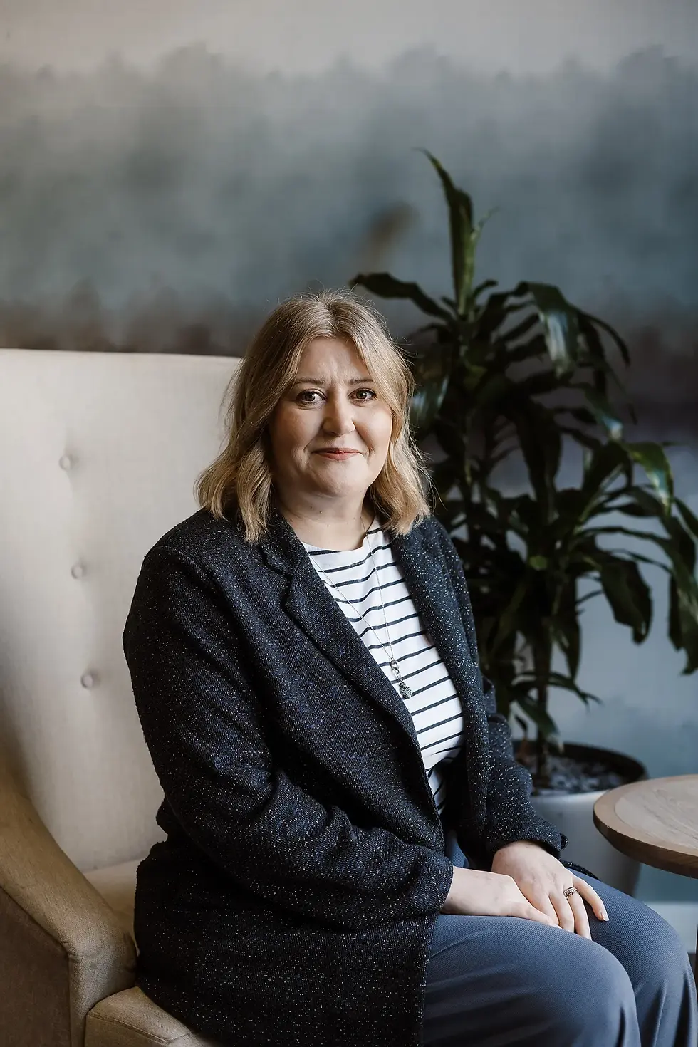 Professional headshot of Danni Foster sitting in a large cream armchair, wearing a dark tweed blazer over a black and white striped top.
