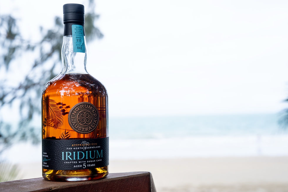 A bottle of FNQ Rum Co. Iridium 5yr Dark Rum standing on a timber rail above the sea